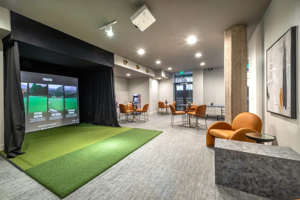 Clubhouse area with a large golf simulator with a green, several chairs at tables, a ping pong table, and 2 arcade machines