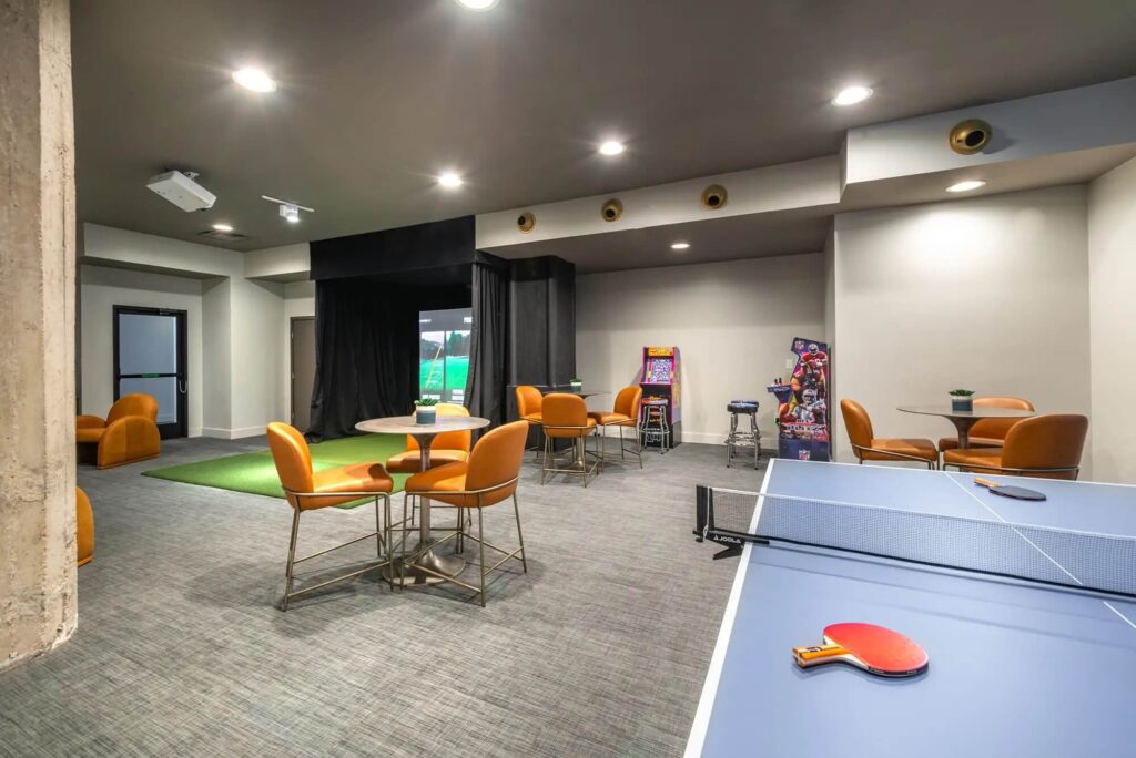 Clubhouse area with a large golf simulator with a green, several chairs at tables, a ping pong table, and 2 arcade machines, fun lighting
