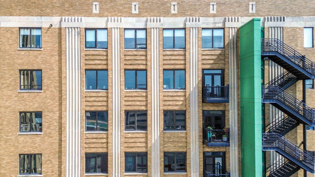 A tan brick building facade with vertical stone pilasters, grid windows, and an external black metal staircase attached to a bright green side section.