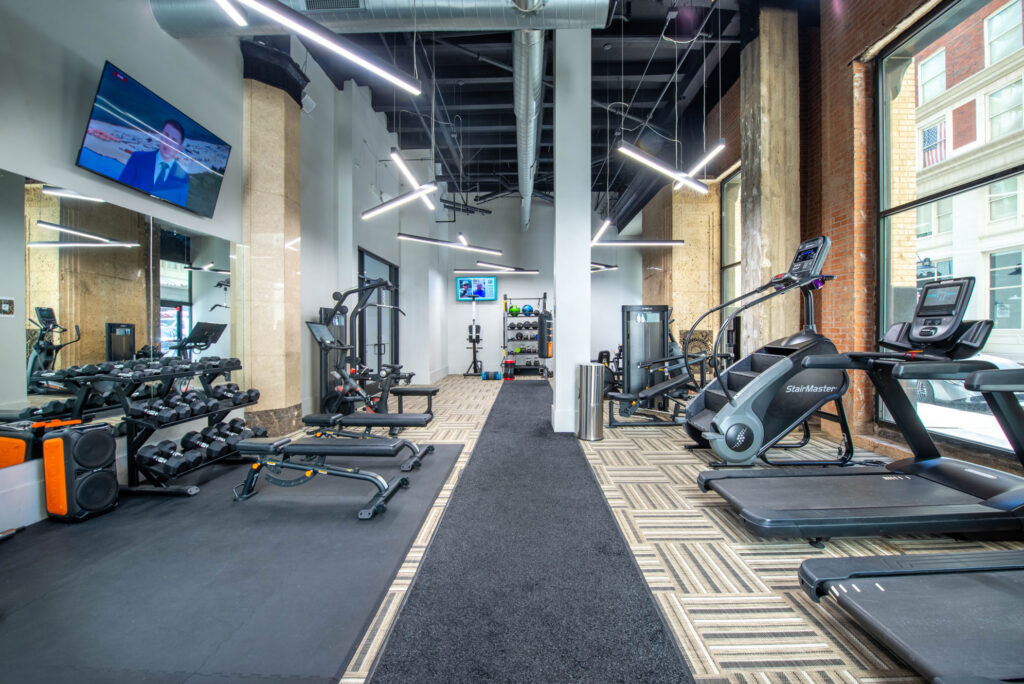 A gym interior featuring a central aisle with weight benches, a dumbbell rack, a StairMaster, and treadmills, with a television mounted on the wall.