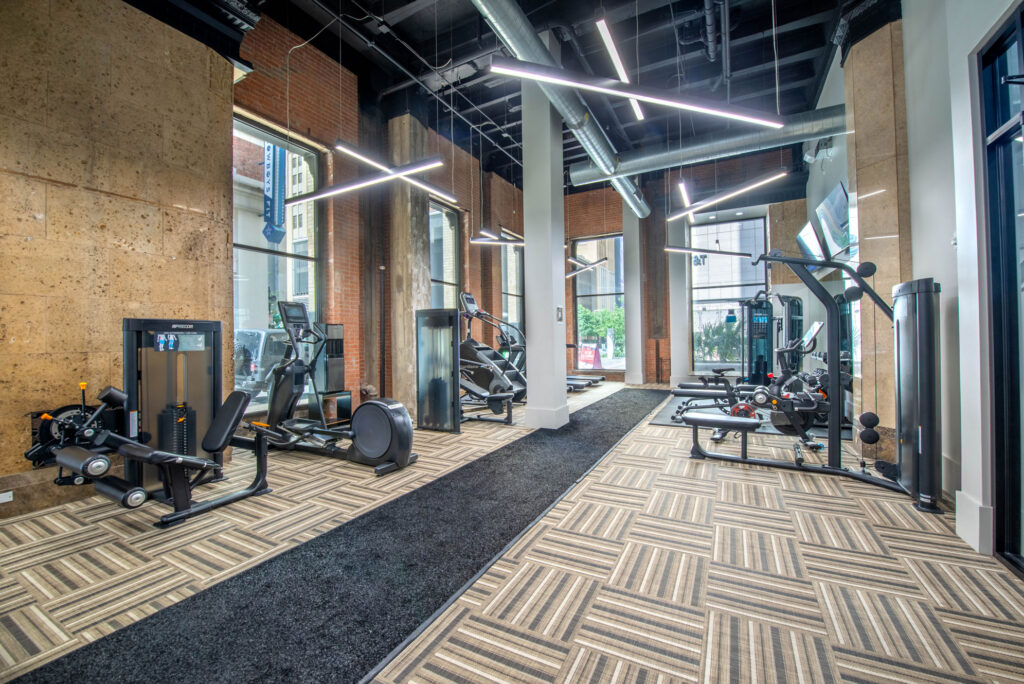 A gym interior with various fitness machines including weight stations and elliptical trainers arranged on a patterned carpeted floor.