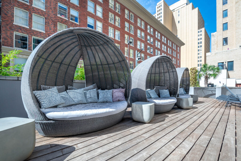 Three rounded, grey wicker daybeds with cushions lined up on a wooden rooftop deck in front of a brick building.