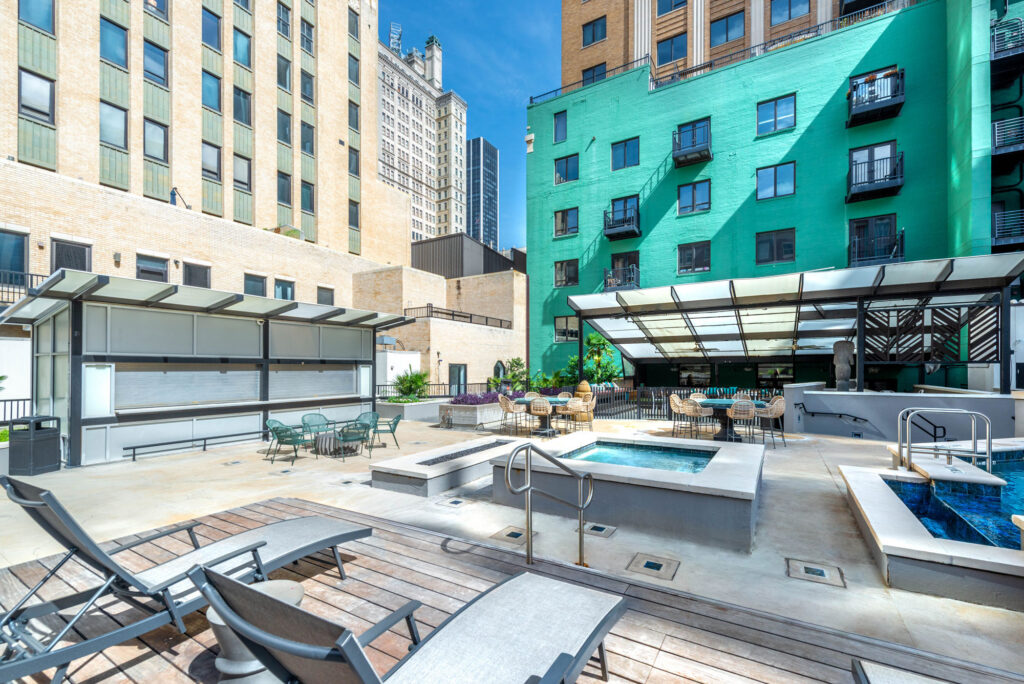 A hotel rooftop terrace with lounge chairs, a hot tub, outdoor dining tables, and a bar area set between tall brick and green buildings under a clear sky.