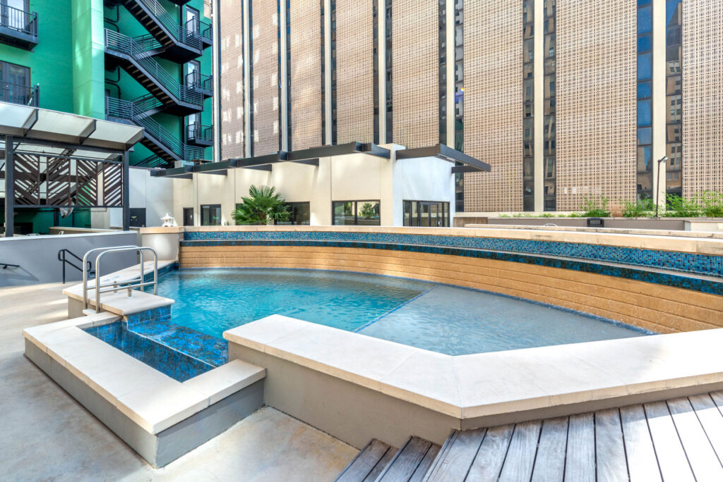 A curved swimming pool with a tiered blue tiled ledge and an adjacent hot tub on a concrete terrace surrounded by tall modern buildings.