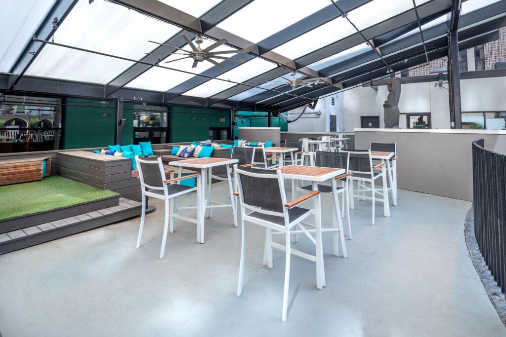 Outdoor patio featuring multiple high-top tables with metal chairs, a tiered wooden platform with blue cushions, and a retractable glass roof.