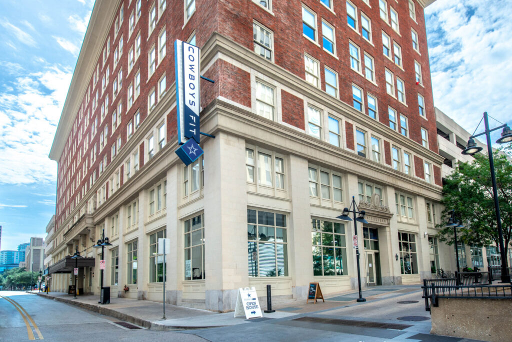 The Cowboys Fit building on a corner street, featuring a vertical blue sign and an Open House sandwich board sign on the sidewalk.