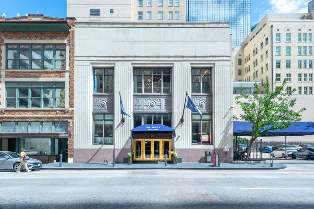 The Joule hotel entrance on Main Street, featuring a blue awning over gold-framed glass doors set into a neoclassical facade.