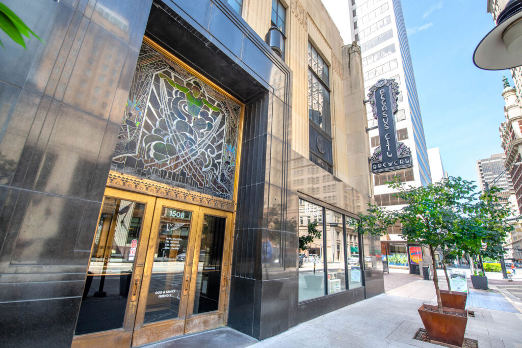 The entrance of the Pegasus City Brewery building, featuring an ornate stained glass transom window above brass-framed doors on a city sidewalk.