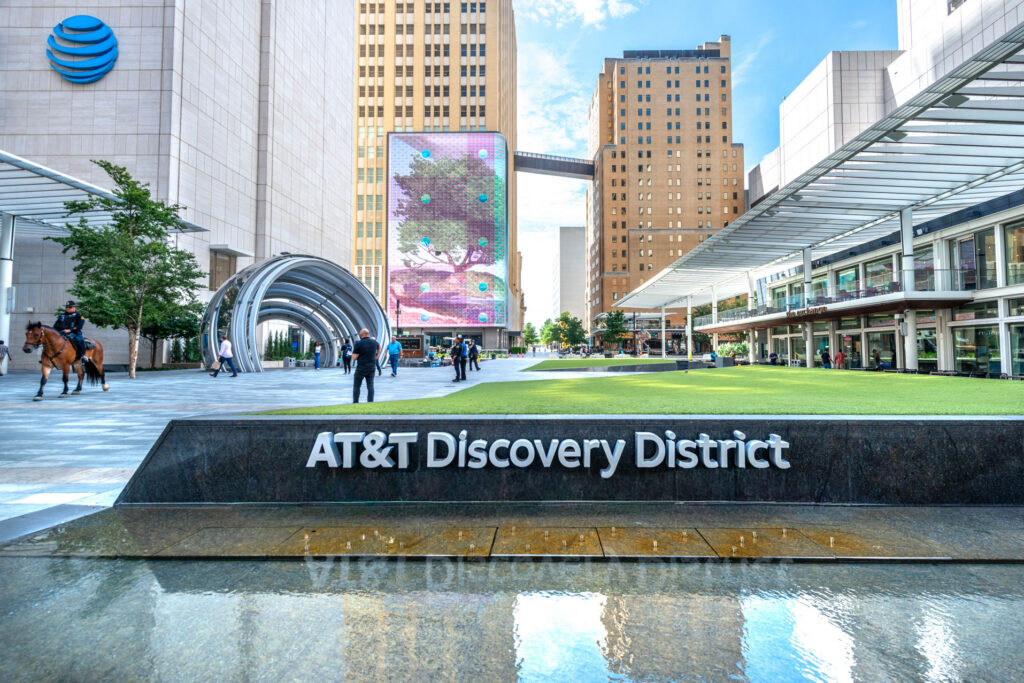 The AT&T Discovery District plaza featuring a large digital display, a metal sculpture, and a mounted police officer, with the company logo on a stone wall in the foreground.