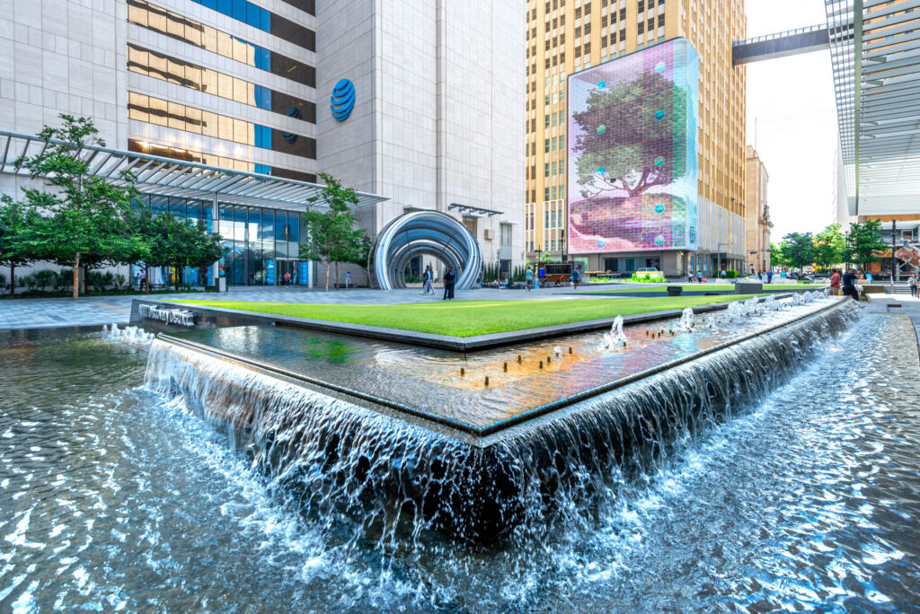 A tiered rectangular fountain with cascading water in a modern urban plaza featuring a large building with the AT&T logo, a metallic sculpture, and a digital billboard of a tree.