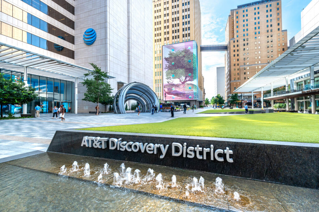 The AT&T Discovery District plaza featuring a large digital display, a sculpture, and a fountain with signage in the foreground.