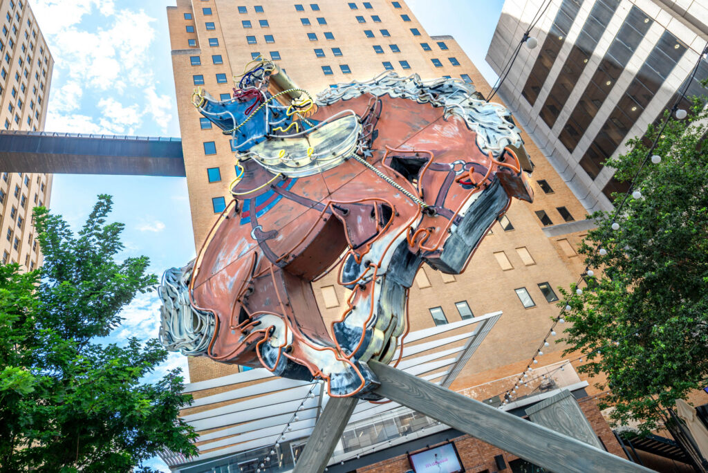A large neon sculpture of a cowboy riding a bucking horse mounted on a wooden frame in front of tall city buildings.
