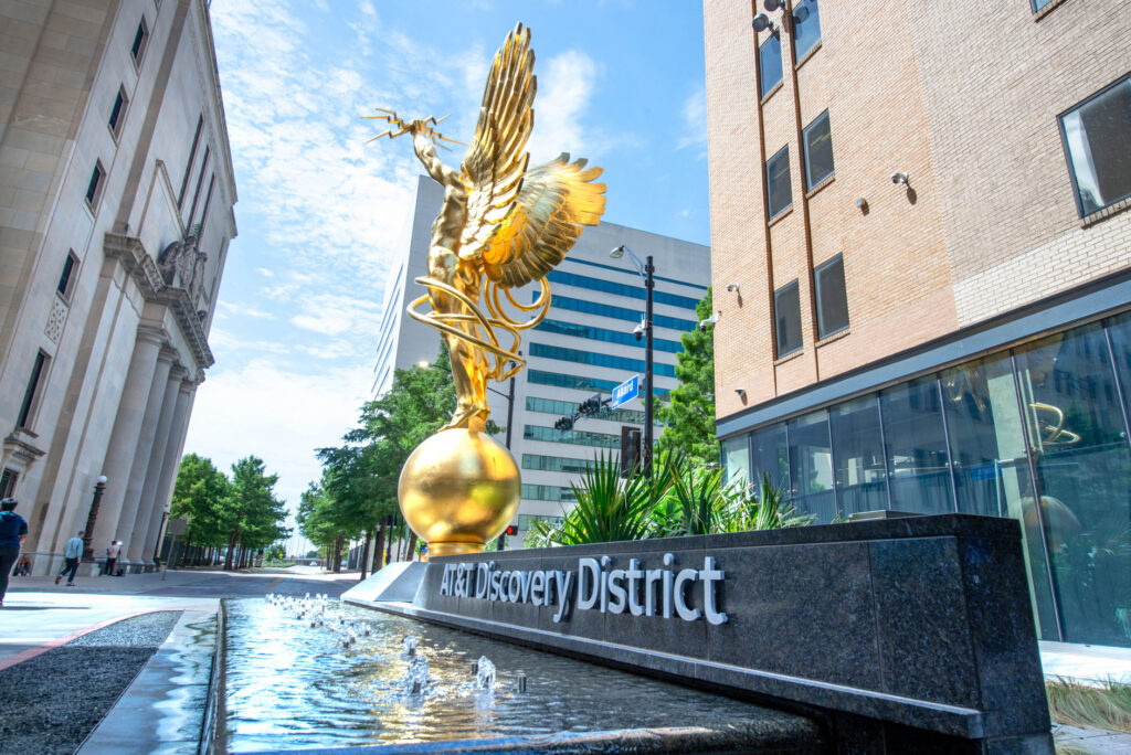 A golden statue of a winged figure holding a lightning bolt atop a sphere at the AT&T Discovery District, featuring a reflecting pool in the foreground.