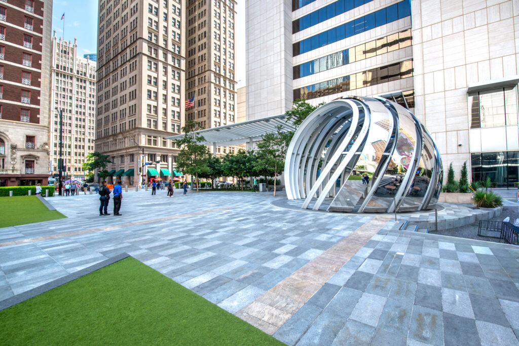 A tiled public plaza featuring a large, curved, silver metal sculpture in front of a tall office building, with several pedestrians walking in the background.
