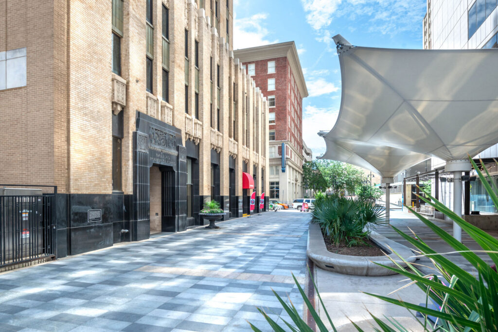 An urban plaza with a checkered stone walkway leading past a tall, beige brick building and a large white architectural shade canopy.