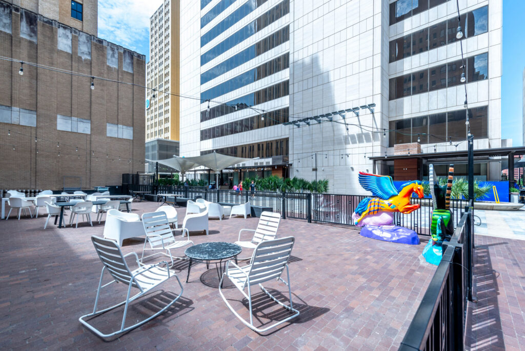 Outdoor patio with white rocking chairs, small tables, and a colorful, winged horse sculpture near a city building.