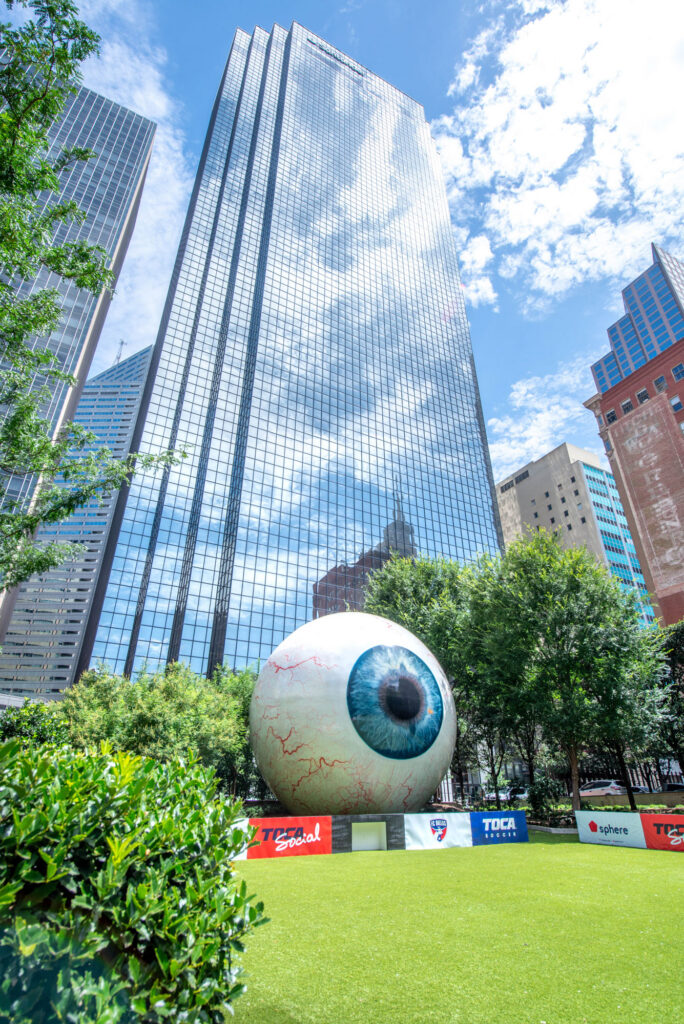 A giant sculpture of a human eyeball sits on a grassy lawn in front of a tall, glass-faced skyscraper.