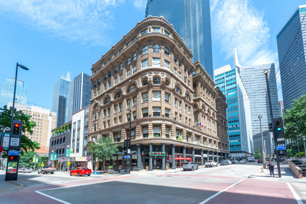 A historic brown stone building with arched windows on a street corner in downtown Dallas, surrounded by modern skyscrapers.