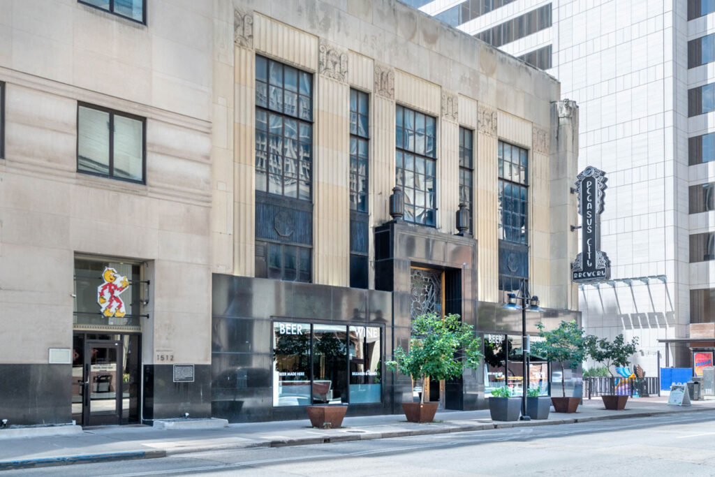 The exterior of the Pegasus City Brewery building, featuring tall windows, a prominent vertical neon sign, and potted trees lining the sidewalk.
