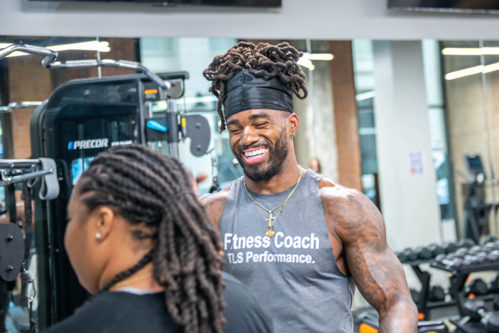 A smiling personal trainer wearing a gray tank top labeled Fitness Coach TLS Performance talks to a client in a gym.