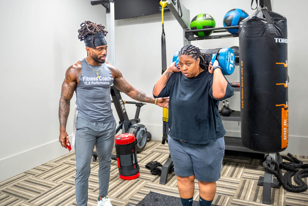 A fitness coach observes as a person performs a weighted squat with a blue sandbag across their shoulders in a gym.