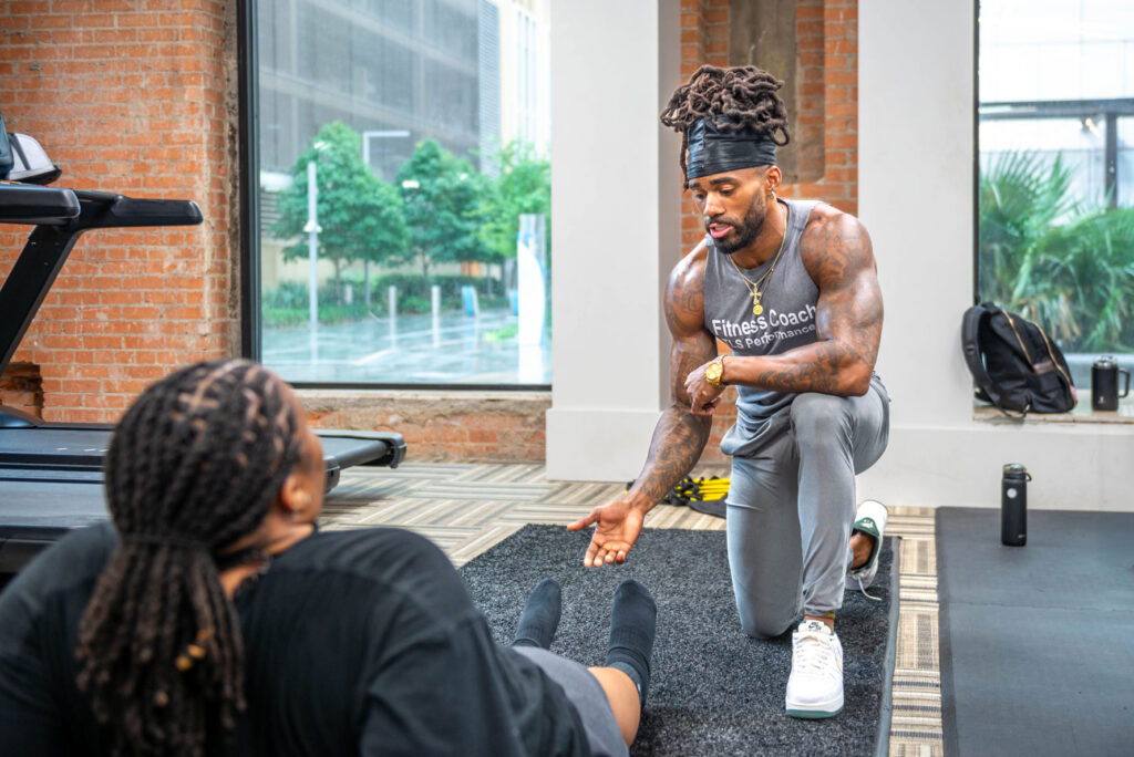 A male fitness coach kneels on a gym mat, instructing a person who is lying on the floor during a workout.