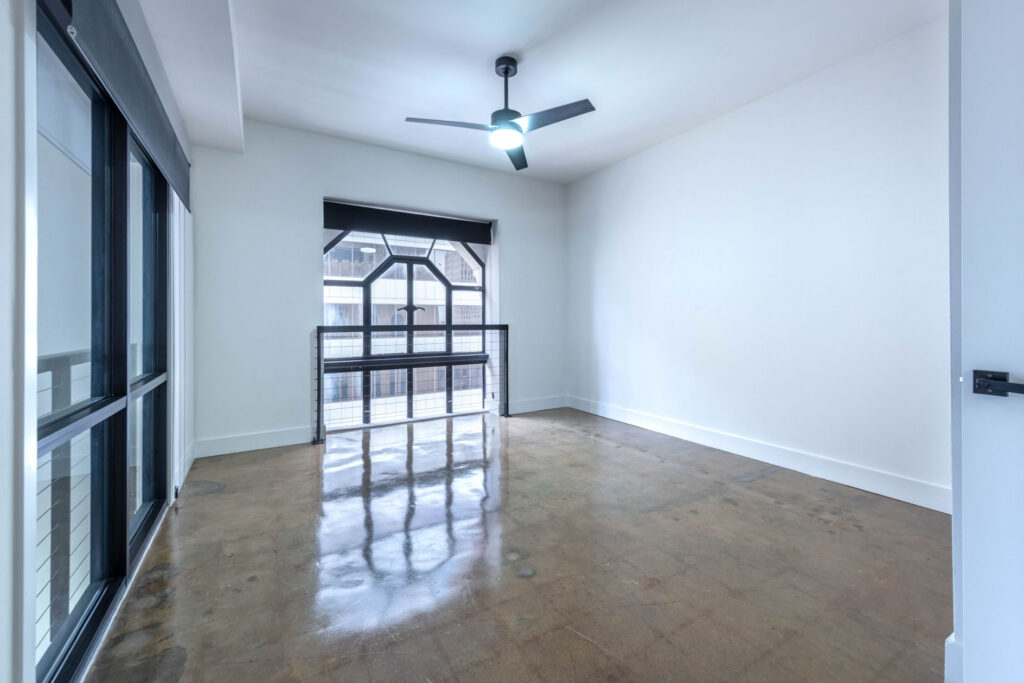 An empty room with white walls, a polished concrete floor, a ceiling fan, and a large window with a black frame and metal railing.