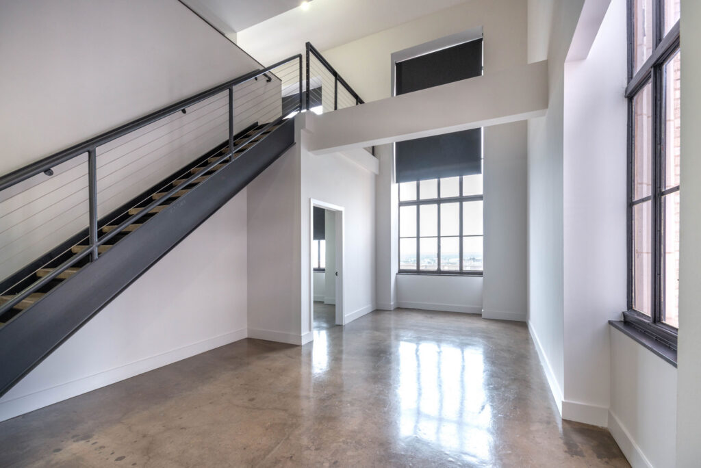 Empty room with polished concrete floors, tall windows, and a metal staircase leading to an upper landing.