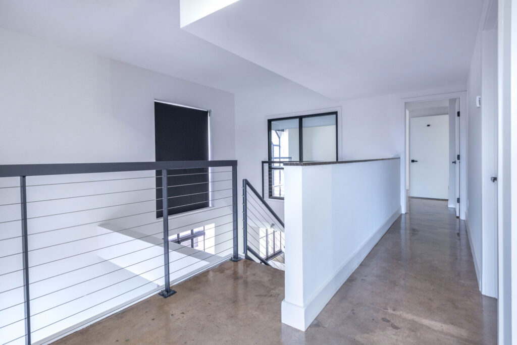 A brightly lit indoor hallway with concrete floors, a metal cable railing overlooking a stairwell, and a doorway leading to another room.