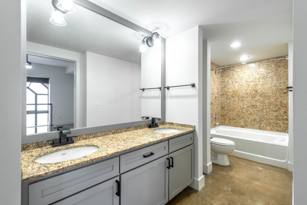 Bathroom with a gray double vanity, granite countertop, two sinks, and a bathtub with a tiled surround.