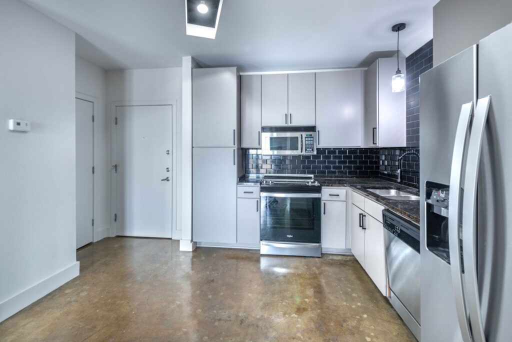 A modern kitchen with light gray cabinets, black tile backsplash, stainless steel appliances, and a polished concrete floor.