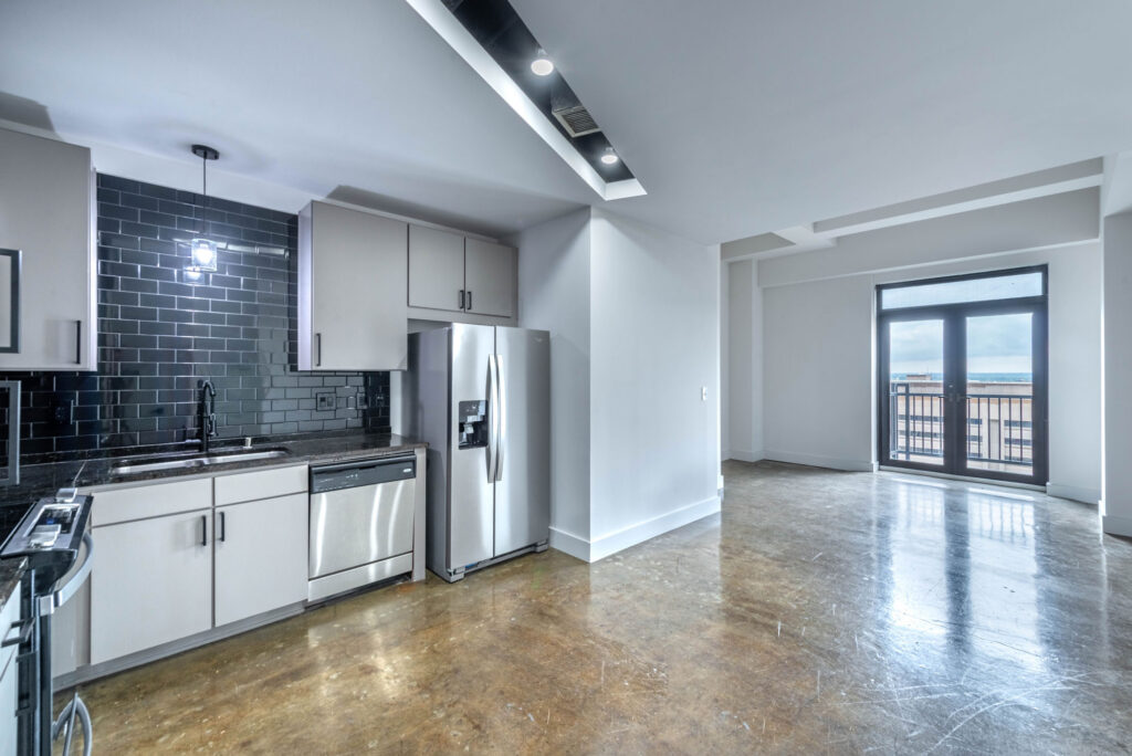 Modern kitchen with stainless steel appliances, black subway tile backsplash, and polished concrete floors, opening into a spacious living area with a balcony door.