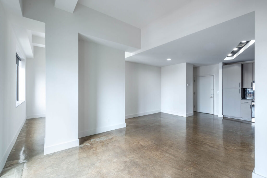 An empty room with white walls, a polished concrete floor, and a small kitchen visible in the background.
