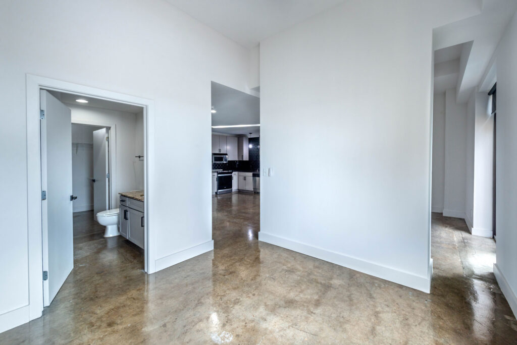 View through an open doorway into a bathroom with a toilet and vanity, with a kitchen visible in the background and a concrete floor in the foreground.