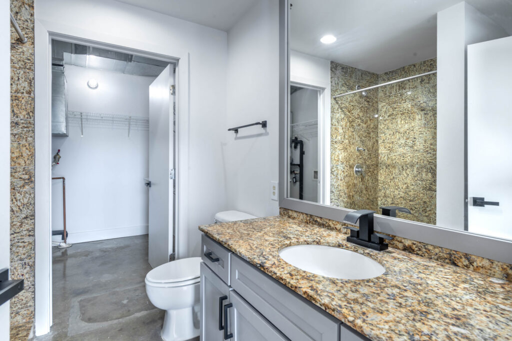 Bathroom with a granite vanity, oval sink, and mirror reflecting a tiled walk-in shower, with an open doorway leading to a walk-in closet.