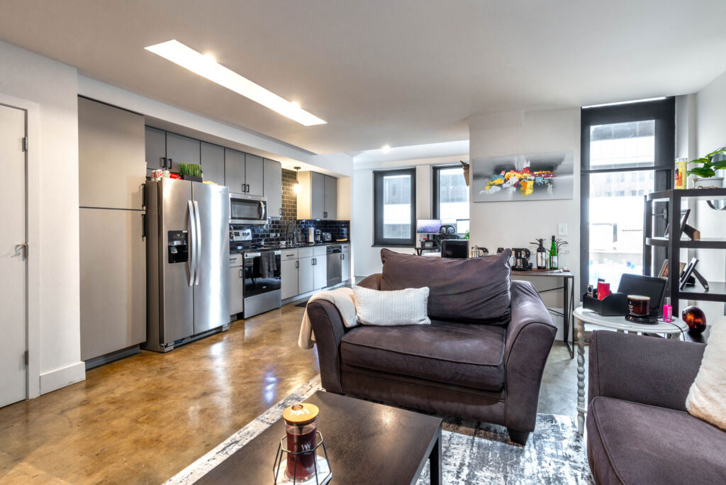A modern open-plan apartment featuring a dark gray sofa in the foreground, a kitchen with stainless steel appliances, and a dining area in the background.