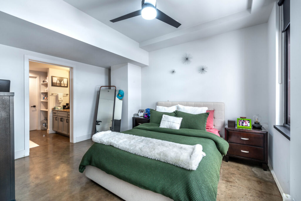 A bedroom with a green bedspread and white faux-fur runner, featuring a dark wood nightstand and a doorway leading to a bathroom.