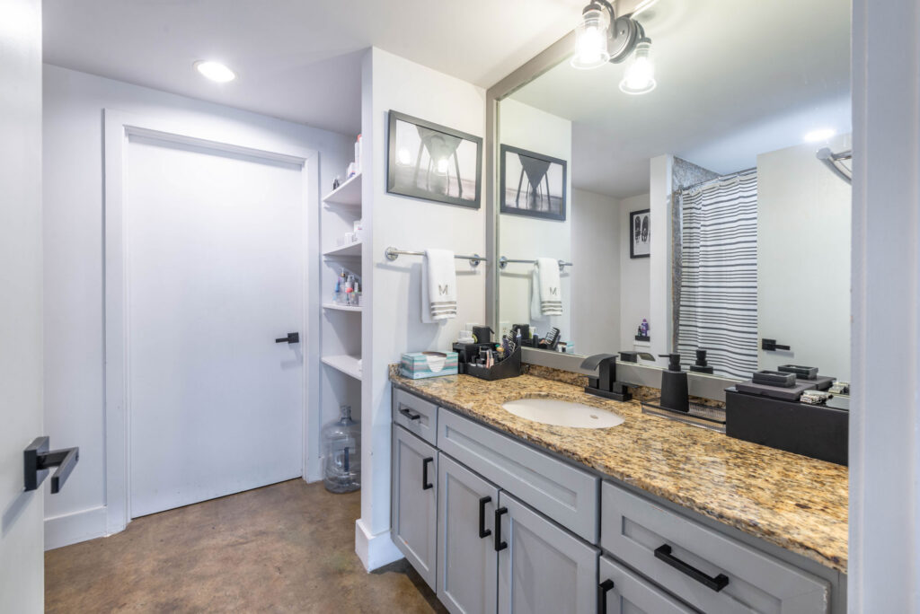 Bathroom vanity with grey cabinets, a speckled stone countertop, and a large mirror, with open shelving and a closed door nearby.