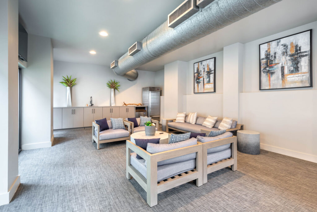 A modern lounge area with gray armchairs and a sofa arranged around a round coffee table, featuring minimalist decor, wall art, and exposed ceiling ductwork.