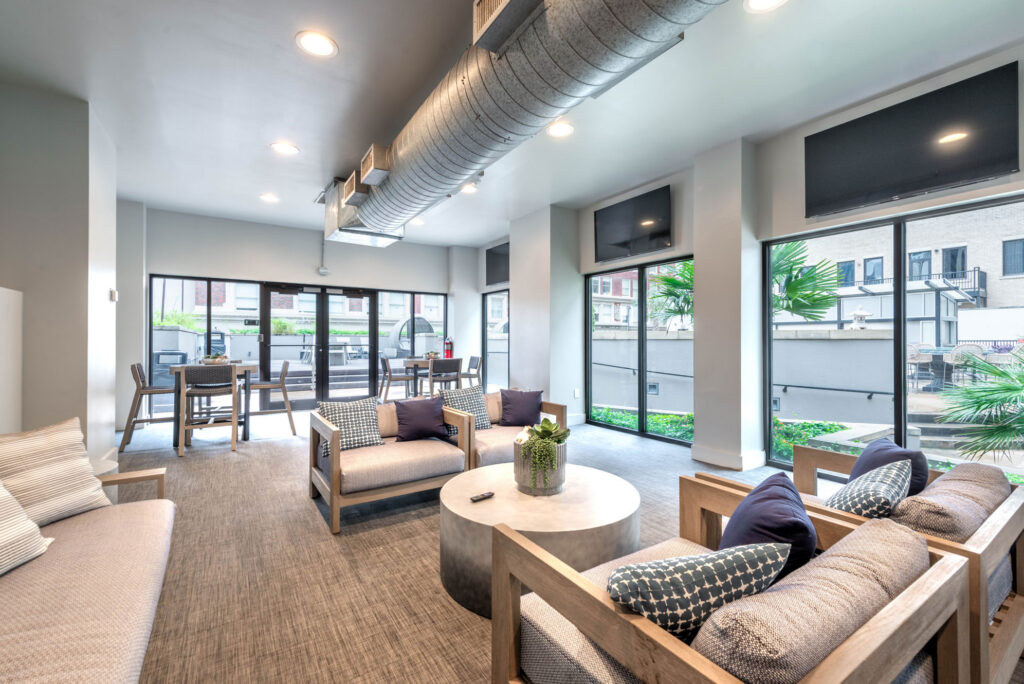 A lounge area with wooden sofas, a round coffee table, and large windows looking out onto an outdoor patio.