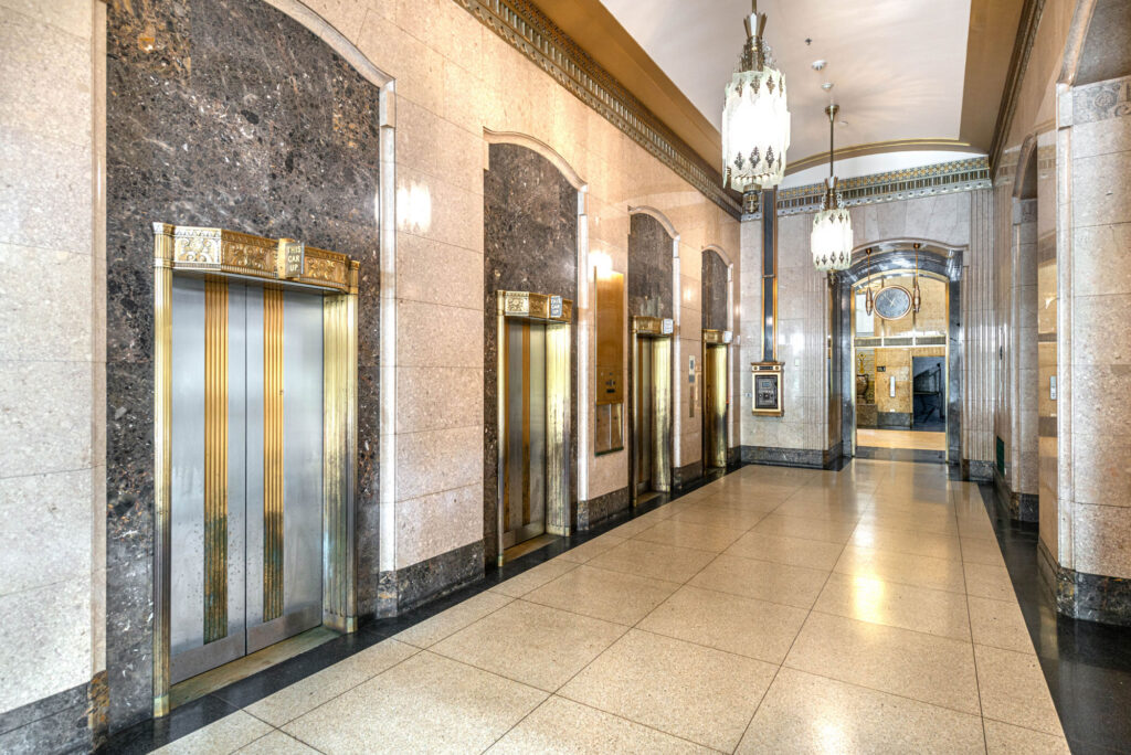 A long hallway featuring a row of ornate art deco elevator doors with gold accents, tile flooring, and hanging glass chandeliers.