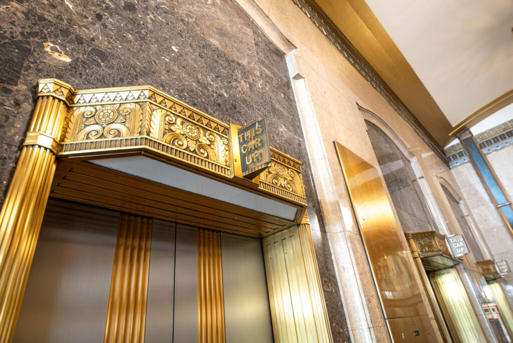 Art deco elevator doors with gold trim and an illuminated sign reading THIS CAR UP in an ornate marble hallway.