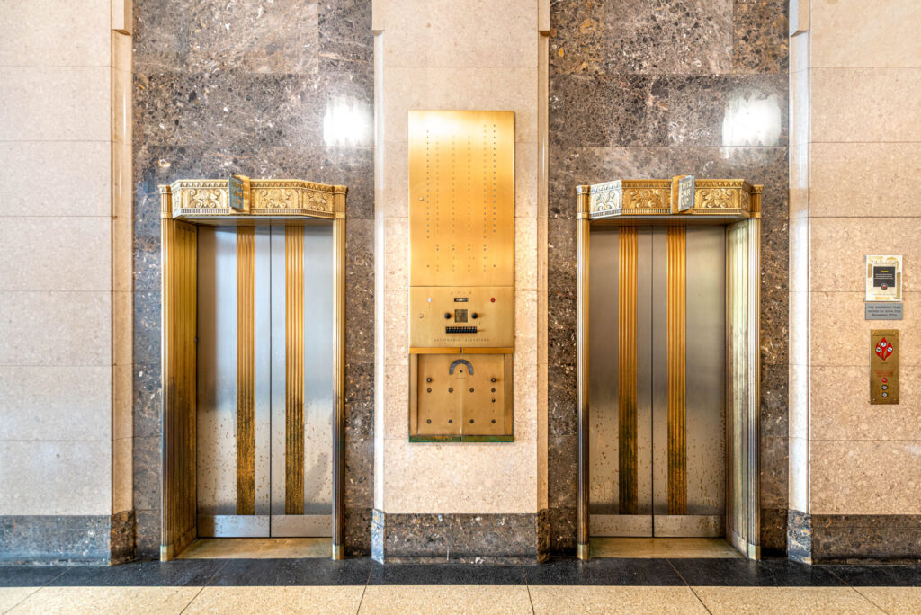 Two elevators with decorative gold-tone metal frames and silver doors, flanking a central brass floor indicator panel set against a marble wall.