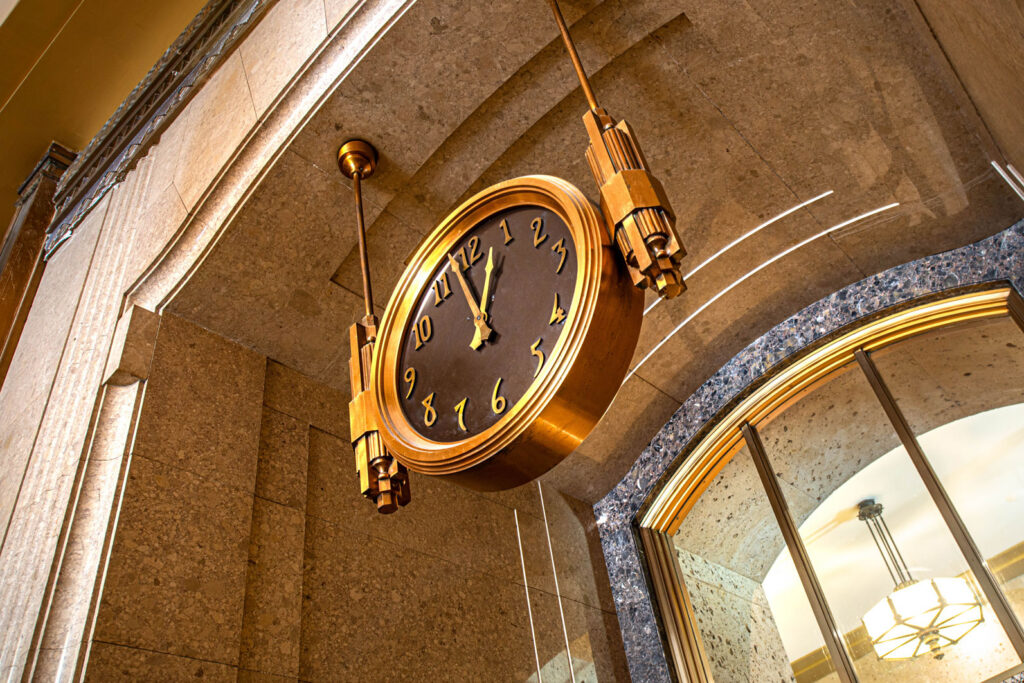 An Art Deco-style, double-faced round clock suspended from a stone ceiling.