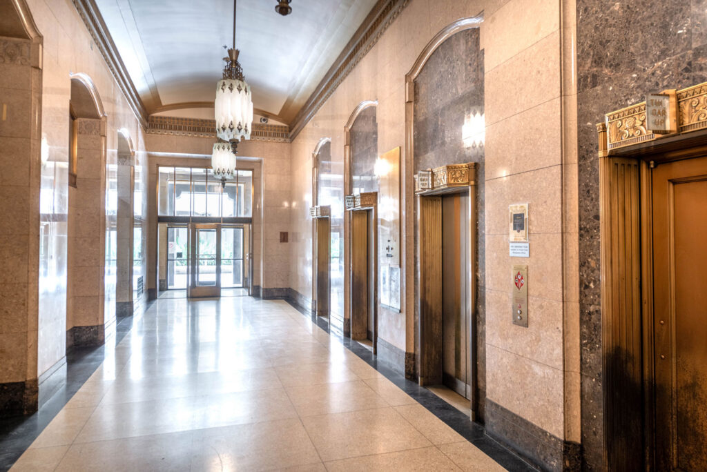 A long hallway with polished stone floors and walls, featuring a series of ornate brass elevator doors and a glass-paneled entrance at the far end.