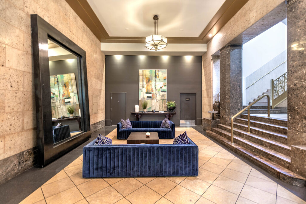 A hotel lobby with two navy blue velvet sofas facing a low coffee table, a large mirror on the left wall, and a staircase on the right.