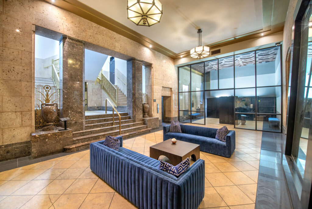 A lobby area with two blue velvet sofas, a wooden coffee table, stone walls, and a staircase leading to an upper level.