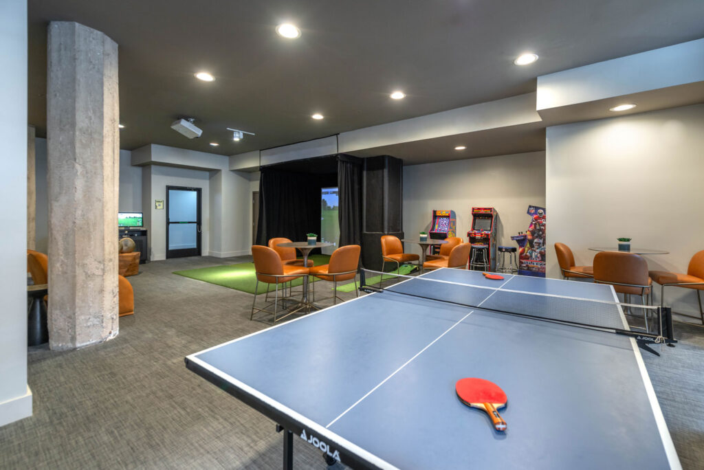 A game room with a blue ping pong table in the foreground, a golf simulator booth, arcade machines, and seating areas with tables and chairs.