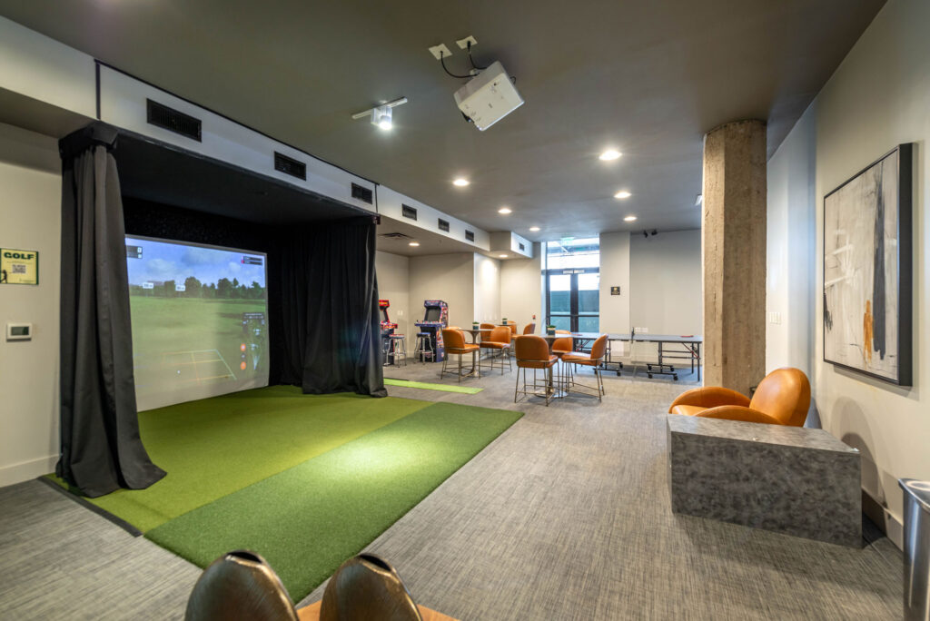 An indoor golf simulator with a putting green area, surrounded by arcade games, tables with orange chairs, and a ping pong table.