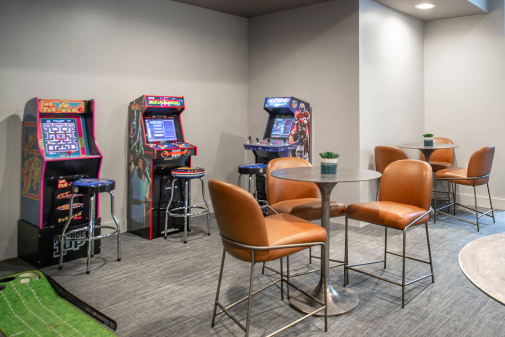 Arcade cabinets for Pac-Man, Mortal Kombat, and NFL Blitz lined up next to caf&eacute;-style tables with brown leather chairs.